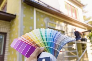 person holding paint samples up to the exterior of a house